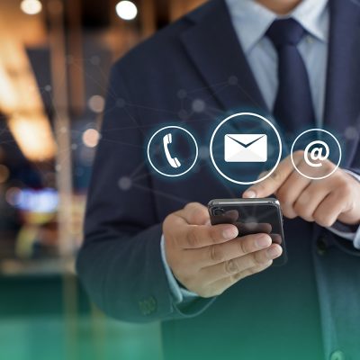 Homem branco de terno e gravata com um celular na mão. Na tela, vemos os ícones de e-mail, mensagem e texto e telefone, meios de comunicação usados em técnicas de vendas.