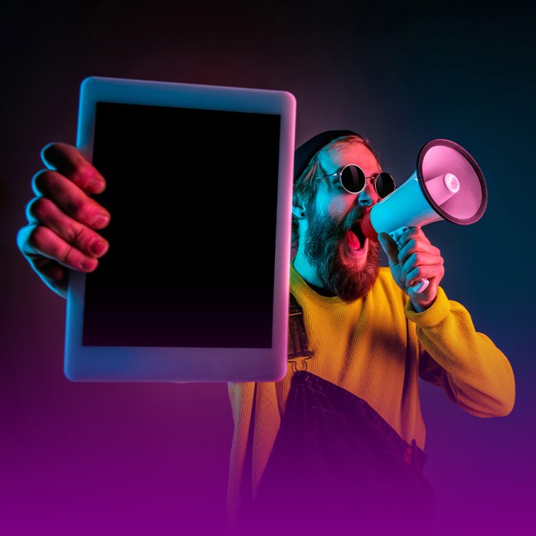 Homem de óculos escuros redondo, barba comprida, usando uma blusa amarela e macacão azul, em fundo escuro com degradê de roxo, fala em um megafone. Na outra mão, em primeiro plano, ele segura um tablet. A imagem representa o alcance do tráfego pago.