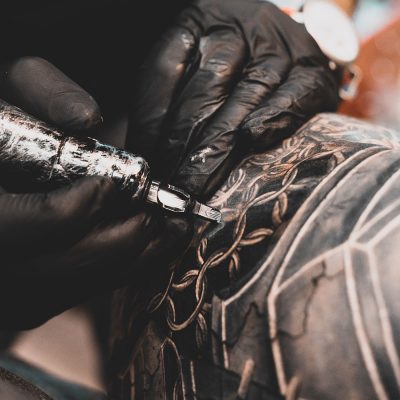 Um par de mãos usando luvas de procedimento pretas segura uma máquina de tatuagem. Ela está tatuando uma pessoa, o desenho tem fundo preto e lembra argolas.
