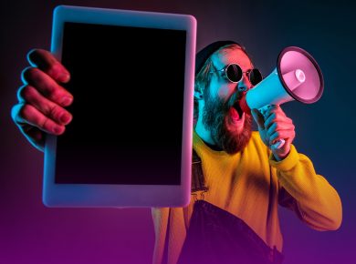 Homem de óculos escuros redondo, barba comprida, usando uma blusa amarela e macacão azul, em fundo escuro com degradê de roxo, fala em um megafone. Na outra mão, em primeiro plano, ele segura um tablet. A imagem representa o alcance do tráfego pago.