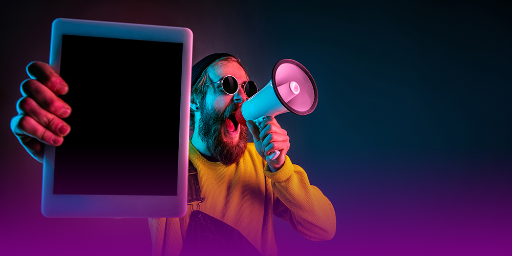 Homem de óculos escuros redondo, barba comprida, usando uma blusa amarela e macacão azul, em fundo escuro com degradê de roxo, fala em um megafone. Na outra mão, em primeiro plano, ele segura um tablet. A imagem representa o alcance do tráfego pago.