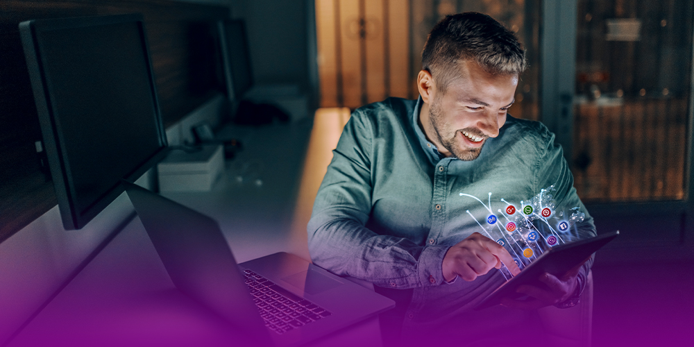 Um homem está sorrindo segurando um tablet com diversos ícones de redes sociais (instagram, facebook e outras, onde acontece a maior parte do tráfego orgânico) "saindo" da tela. Ele está sentado em frente a um notebook e a uma tela desligada.