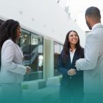 Três pessoas estão em pé em uma sala ampla, como um saguão de uma empresa. Todos estão vestindo roupas formais. Um homem e uma mulher ligeiramente de costas, ambos negros e vestindo paletó cinza-claro, estão sorrindo e cumprimentando uma terceira mulher. Ela é branca, está com as mãos cruzadas de forma amigável na altura da cintura e sorri. Também notamos o rapport pela linguagem corporal deles.
